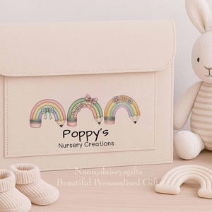 May include: A beige felt nursery clutch with a flap closure, featuring a colorful rainbow pencil design and the text "Poppy's Nursery Creations." A stuffed bunny, baby booties, and a rainbow-shaped toy are also present.