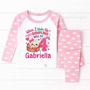 May include: A long-sleeved pyjama set featuring a white top with pink raglan sleeves and pink trousers, both patterned with white clouds. The top displays a graphic of a cartoon dumpling in a steamer, the number 4, and the text "When I Wake Up This Dumpling Will Be 4 Gabriella."