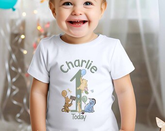 Puede incluir: Camiseta blanca de manga corta con un número uno verde, un personaje de Winnie de Pooh sosteniendo un globo azul y el texto "Charlie" y "Hoy".