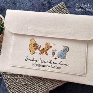 May include: A white felt folder with a Winnie the Pooh design. The folder has the text "Baby Wickenden Pregnancy Notes" printed on it.