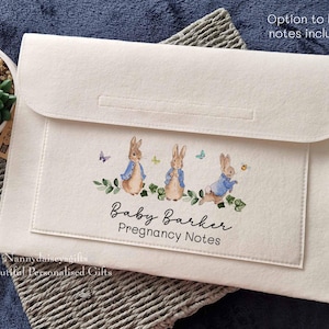 May include: A white felt folder with a flap closure. The folder has a design of three Peter Rabbit characters with the text "Baby Barker Pregnancy Notes".