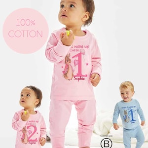 May include: Pink and blue long-sleeved pyjamas for a baby with a Peter Rabbit design. The pyjamas feature a pink bunny with a pink heart and the text "I wake up I will be 1 Sophie". The pyjamas are made of 100% cotton.