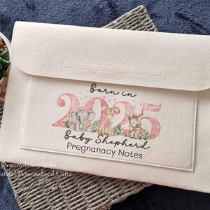 May include: A white felt laptop sleeve with a pink and white design. The design features the text "Born in 2025 Baby Shepherd Pregnancy Notes".