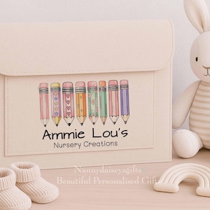 May include: A beige felt folder with a flap, decorated with a pencil design and the text "Ammie Lou's Nursery Creations." A stuffed bunny, baby booties, and a rainbow-shaped toy are also visible.