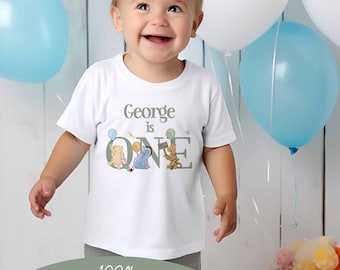 Puede incluir: Una camiseta blanca con un diseño de Winnie the Pooh. La camiseta dice "George is One" con un personaje de Winnie the Pooh sosteniendo un globo. La camiseta es 100% algodón suave.