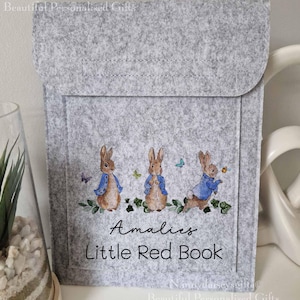 May include: A grey felt book cover with a personalised name "Amales" and the text "Little Red Book". The cover features three illustrated Peter Rabbit characters in blue jackets.