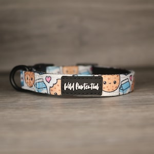 May include: A white dog collar with a colourful pattern of cookies and milk cartons. The collar has a black buckle and a black tag that says "Wild Paw-tential".
