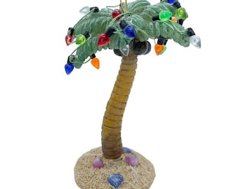 Puede incluir: Un adorno decorativo de palmera con un tronco marrón texturizado y frondas verdes adornadas con pequeñas bombillas multicolores en rojo, azul, verde, naranja y blanco. El árbol se asienta sobre una base arenosa decorada con conchas marinas.
