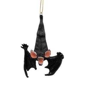 May include: A black hanging bat ornament with a textured body, large pinkish-tan ears, and an open mouth revealing white teeth. The bat is suspended by a thin gold string attached to its feet, with its wings spread wide in a downward pose.
