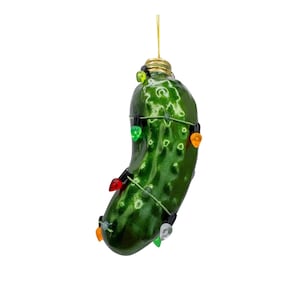 May include: A green pickle-shaped Christmas ornament with a gold cap and a string of colorful lights wrapped around it. The ornament hangs from a gold string against a white background.
