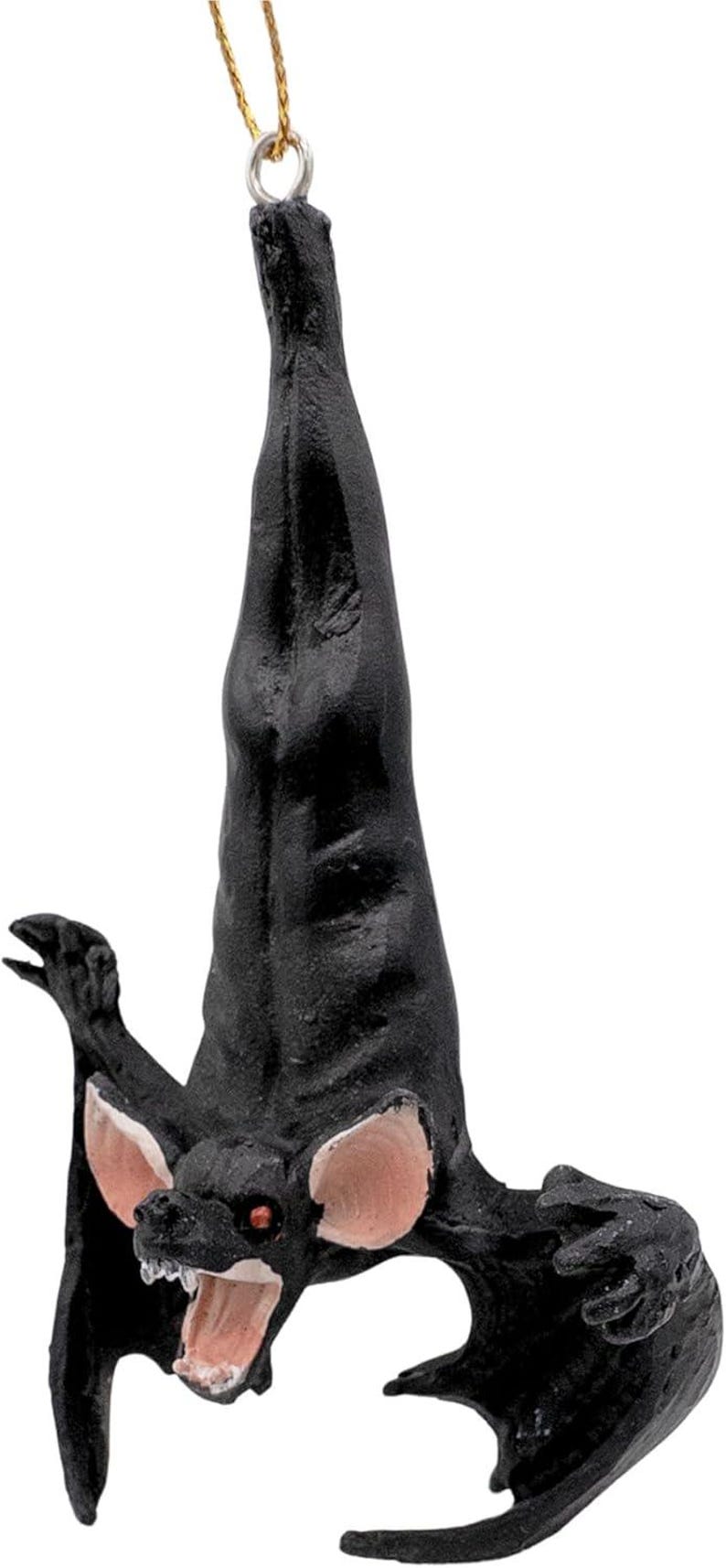 May include: A black bat ornament hanging upside down by a gold string. The bat has large, light pink ears, red eyes, and an open mouth revealing white teeth. Its wings are partially spread, and its body features a textured, matte black finish.