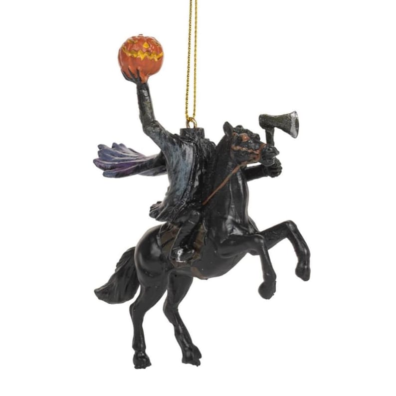 Puede incluir: Un adorno colgante que representa a un jinete sin cabeza con abrigo oscuro y capa morada, montado sobre un caballo negro. El jinete sostiene una calabaza naranja tallada en alto con una mano y un hacha en la otra. Incluye un cord&oacute;n dorado.