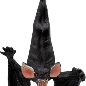 May include: A black hanging bat ornament with a textured body, large pink-lined ears, and an open mouth showing white teeth. The bat is suspended by a thin gold string attached to a metal loop at the top of its head, featuring a dark, detailed design.