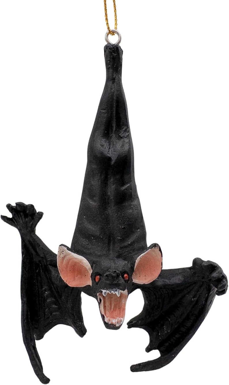 May include: A black hanging bat ornament with a textured body, large pink-lined ears, and an open mouth showing white teeth. The bat is suspended by a thin gold string attached to a metal loop at the top of its head, featuring a dark, detailed design.