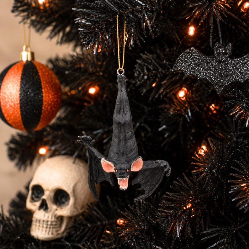 May include: A hanging bat ornament featuring a black body, outstretched wings, pink inner ears, and a mouth open to reveal white teeth and red eyes. The bat is suspended by a thin gold string, showcasing a detailed texture and a downward-facing pose.
