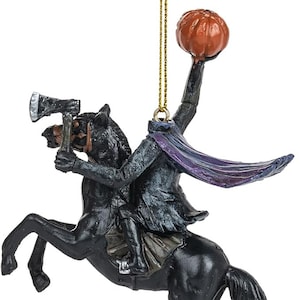 Puede incluir: Un adorno colgante que representa a un jinete sin cabeza sobre un caballo negro. El jinete viste un abrigo oscuro con una capa morada, sostiene un hacha en una mano y levanta una calabaza naranja sobre su cuello. Un cord&oacute;n dorado sirve para colgarlo.