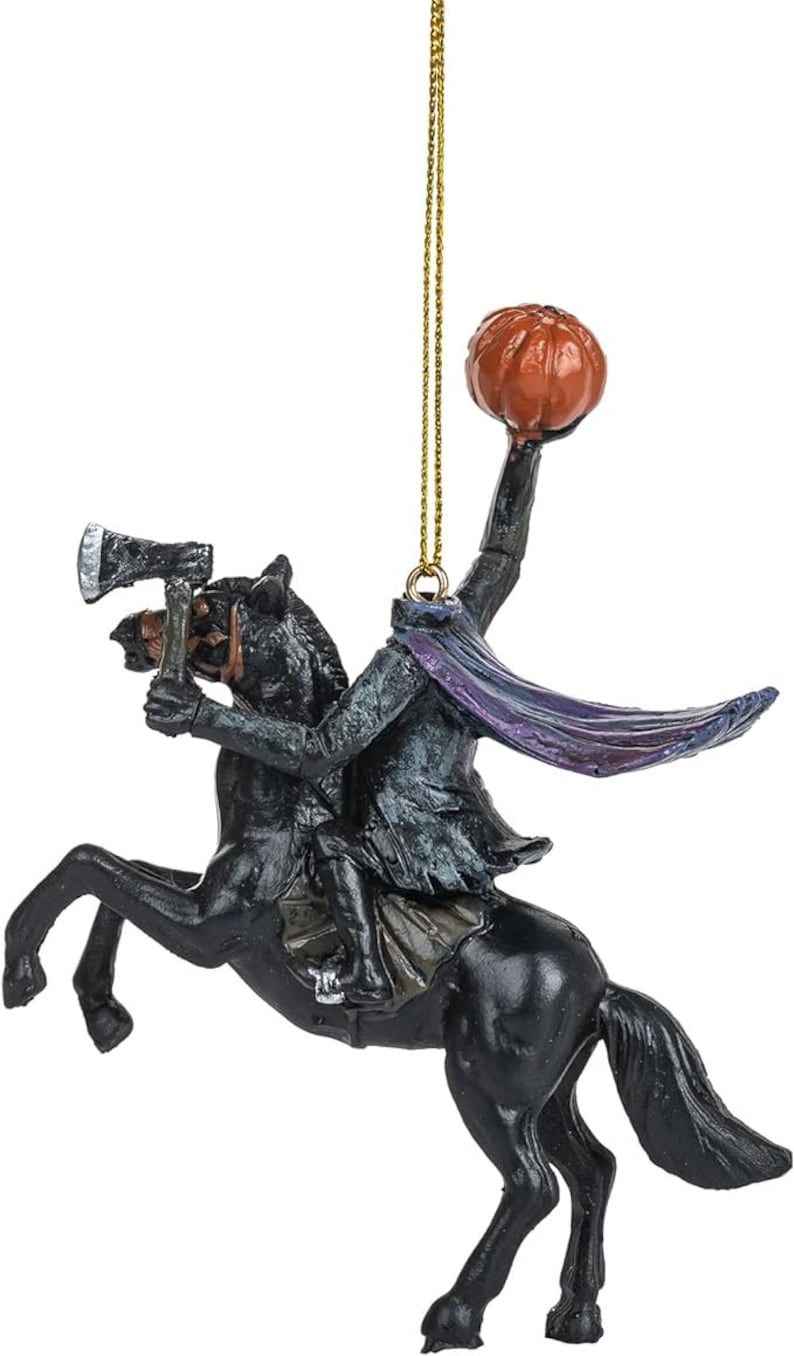 Puede incluir: Un adorno colgante que representa a un jinete sin cabeza sobre un caballo negro. El jinete viste un abrigo oscuro con una capa morada, sostiene un hacha en una mano y levanta una calabaza naranja sobre su cuello. Un cord&oacute;n dorado sirve para colgarlo.