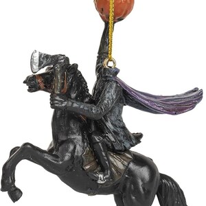 Puede incluir: Un adorno colgante que representa a un jinete sin cabeza con abrigo oscuro y capa morada, montado sobre un caballo negro encabritado. La figura sostiene un hacha plateada y levanta una calabaza naranja tallada sobre su cuello.