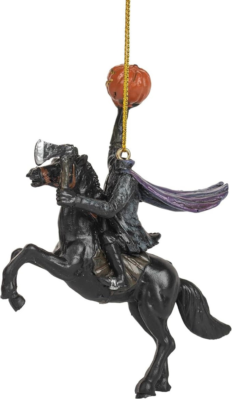 Puede incluir: Un adorno colgante que representa a un jinete sin cabeza con abrigo oscuro y capa morada, montado sobre un caballo negro encabritado. La figura sostiene un hacha plateada y levanta una calabaza naranja tallada sobre su cuello.