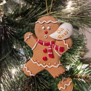 May include: A gingerbread man Christmas ornament with a surprised expression and a speech bubble that says "Oh Snap!" The gingerbread man is wearing a red and white striped scarf and has red buttons.