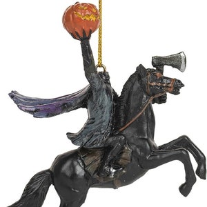 Puede incluir: Un adorno colgante que representa a un jinete sin cabeza con abrigo oscuro y capa forrada de morado, montado sobre un caballo negro encabritado. El jinete sostiene una calabaza naranja tallada en alto, con un hacha plateada en la cabeza del caballo.