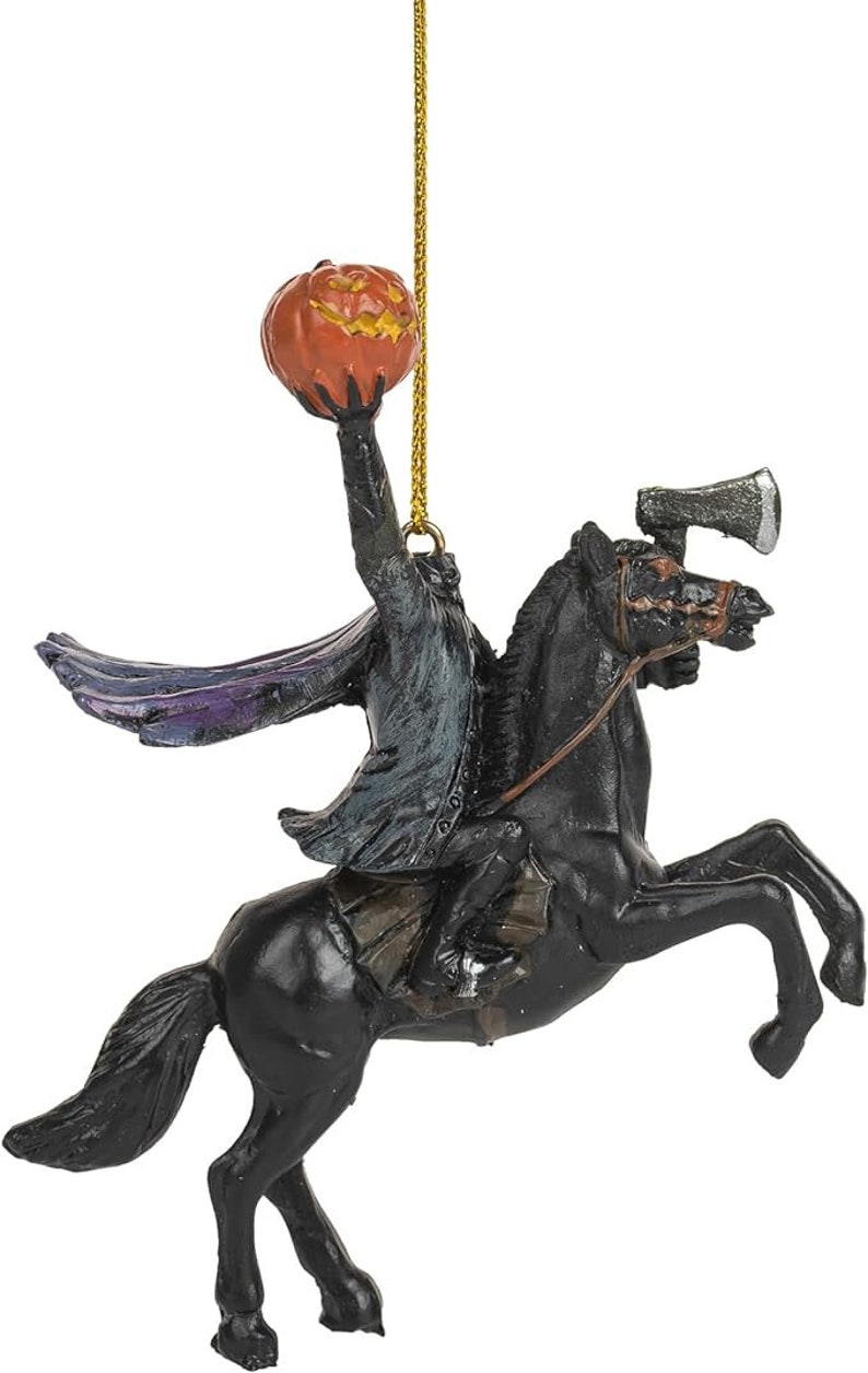 Puede incluir: Un adorno colgante que representa a un jinete sin cabeza con abrigo oscuro y capa forrada de morado, montado sobre un caballo negro encabritado. El jinete sostiene una calabaza naranja tallada en alto, con un hacha plateada en la cabeza del caballo.