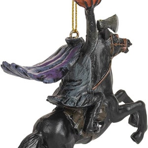 Puede incluir: Un adorno colgante que representa a un jinete sin cabeza sobre un caballo negro. El jinete viste un abrigo oscuro con reflejos morados y sostiene una calabaza naranja en alto. Un hacha es visible sobre el lomo del caballo.