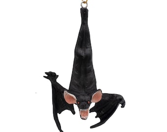 May include: A hanging ornament shaped like a black bat with its wings partially spread. The bat is depicted in an upside-down position with its head facing forward, featuring pink inner ears, red eyes, and an open mouth showing white teeth.