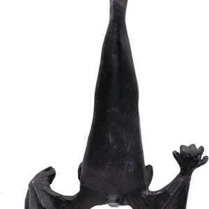 May include: A black hanging ornament shaped like a bat suspended upside down by a thin gold cord. The bat is depicted with its wings partially spread and folded, showing a textured surface and a detailed form with pointed ears and clawed wing tips.