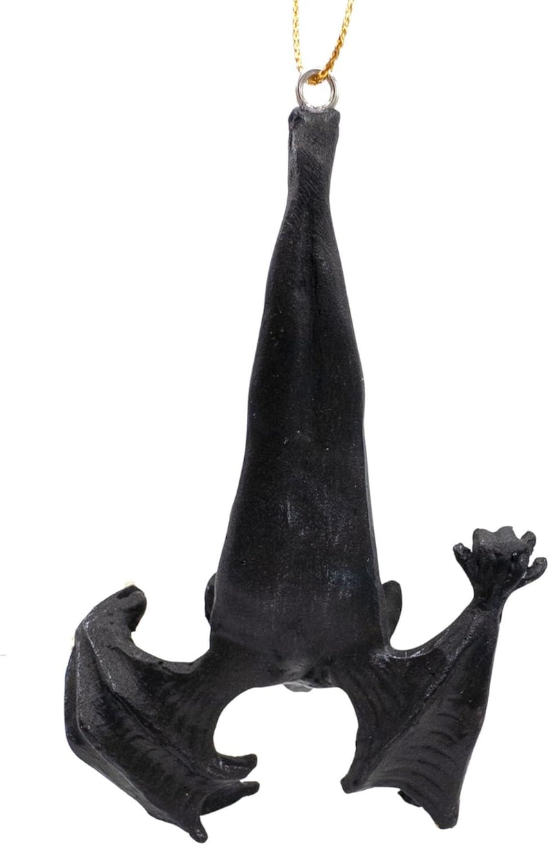 May include: A black hanging ornament shaped like a bat suspended upside down by a thin gold cord. The bat is depicted with its wings partially spread and folded, showing a textured surface and a detailed form with pointed ears and clawed wing tips.