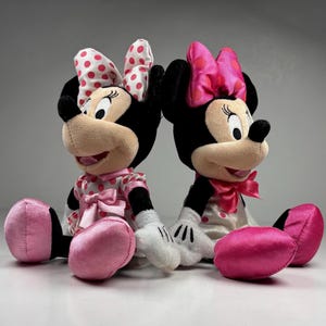 Op de afbeelding: Twee pluche Minnie Mouse poppen. Elke pop heeft zwarte oren, een bruin gezicht en een grote strik met roze stippen. Ze dragen jurken met roze stippen en roze strikken. Hun voeten zijn roze.
