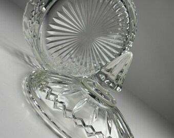 Op de afbeelding: Een heldere glazen bonbonschaal met een deksel. De schaal heeft een ronde basis met een stervormig patroon en een decoratieve rand. Het deksel heeft een ruitpatroon en een puntige knop.