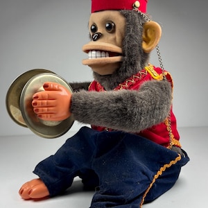 May include: Vintage wind-up toy monkey in a red jacket and blue pants, playing cymbals. The monkey wears a red hat with gold trim and has a chain attached to its ear. The toy's hands and feet are orange, and its fur is gray.
