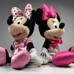 Op de afbeelding: Twee Minnie Mouse pluche poppen. De ene pop draagt een roze en witte polkadot jurk en strik, de andere draagt een roze strik en een witte jurk met roze stippen. Beide hebben zwarte oren, neuzen en lachende gezichten.
