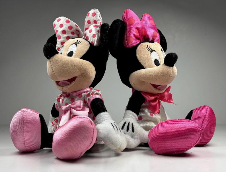 Op de afbeelding: Twee Minnie Mouse pluche poppen. De ene pop draagt een roze en witte polkadot jurk en strik, de andere draagt een roze strik en een witte jurk met roze stippen. Beide hebben zwarte oren, neuzen en lachende gezichten.