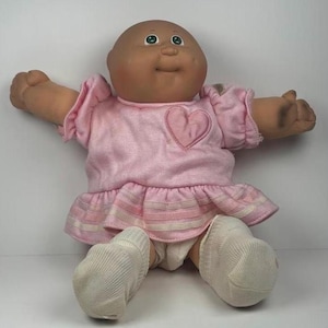 Può includere: Bambola vintage Cabbage Patch Kid con testa calva, occhi verdi e espressione sorridente. La bambola indossa un vestito rosa con un cuore applicato e calzini bianchi. Le braccia della bambola sono tese.