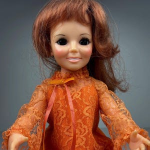 Peut inclure: Poupée vintage aux cheveux roux et au sourire. Elle porte une robe en dentelle orange à manches longues et col montant, ornée d'un ruban pêche. Le visage de la poupée a des joues roses et de grands yeux sombres.