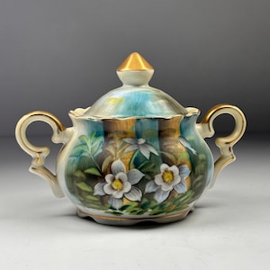 Pode incluir: Uma açucareiro de porcelana vintage com tampa, com um design floral. O açucareiro tem formato arredondado, duas alças e um adorno dourado na tampa. O padrão floral inclui flores brancas com centros amarelos e folhas verdes.