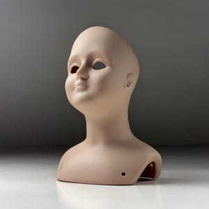 Peut inclure: Une tête de poupée beige avec des orbites oculaires ouvertes, un petit nez et une bouche fermée. Le cou a une forme arrondie avec un trou de chaque côté. La tête est posée sur une surface blanche sur un fond gris.