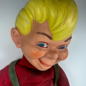 Puede incluir: Una muñeca vintage con una cara sonriente, ojos azules y cabello amarillo. La muñeca lleva una camisa roja y un peto verde. La cara de la muñeca tiene pecas y mejillas rosadas. La muñeca es un objeto de colección.