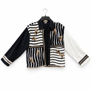 Può includere: Una camicia a maniche lunghe con bottoni dal design color-block, caratterizzata da una manica sinistra nera tinta unita e una manica destra bianca tinta unita. Il pannello centrale presenta strisce verticali bianche e nere con applicazioni triangolari color sabbia.