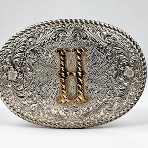 May include: An oval-shaped belt buckle featuring a polished silver-tone finish with intricate scrollwork and floral engravings. A prominent, textured gold-tone letter H is centered on the buckle, which is framed by a twisted rope-style border.