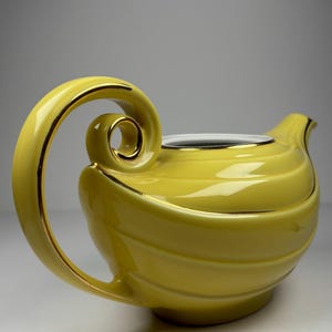 May include: A yellow ceramic teapot with a curved handle and spout. The teapot has gold trim along the handle and rim. The body of the teapot has a ridged design. The teapot is on a white surface.