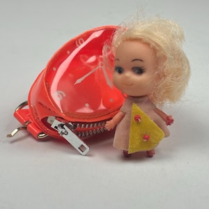 May include: A vintage doll with blonde hair and a pink dress with a yellow triangle accent. Beside the doll is a bright orange, clear plastic purse with a clock face design. The purse has a silver zipper and a small key ring.