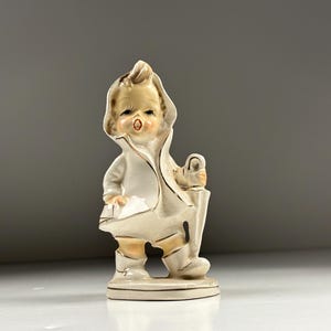May include: A ceramic figurine of a child wearing a hooded raincoat and boots, holding a closed umbrella. The figure is finished in a glossy white glaze with subtle tan shading and delicate gold trim accents along the edges of the clothing.
