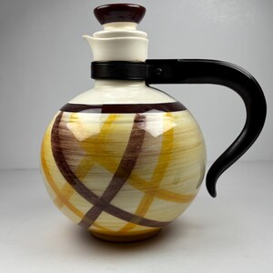 May include: A vintage ceramic pitcher with a round body and a black handle. The pitcher features a cream-colored base with a crisscross pattern in yellow and brown. The lid and handle are black.