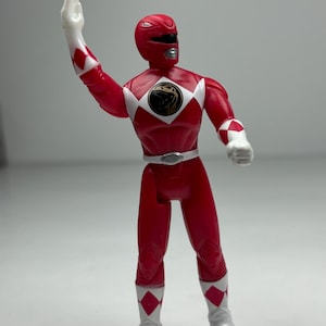 Peut inclure: Figurine rouge de Power Ranger avec des détails blancs, notamment des gants et des bottes. La figurine a une visière noire et un motif de losange blanc sur les épaules et les jambes. Un emblème noir et or est sur la poitrine.