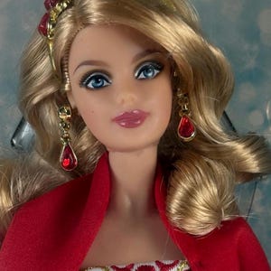 Puede incluir: Una muñeca con cabello rubio, ojos azules y labios rosados, que lleva una diadema dorada con detalles rojos y pendientes a juego en forma de lágrima roja. La muñeca viste una prenda roja sobre un corpiño blanco con un intrincado patrón bordado en rojo y oro.