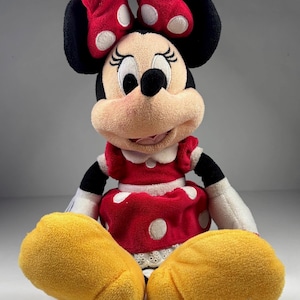 May include: A plush Minnie Mouse doll. The doll has a red dress with white polka dots, a matching bow, and yellow hands. The doll has black ears and a smiling face.