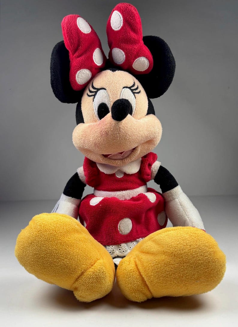 May include: A plush Minnie Mouse doll. The doll has a red dress with white polka dots, a matching bow, and yellow hands. The doll has black ears and a smiling face.